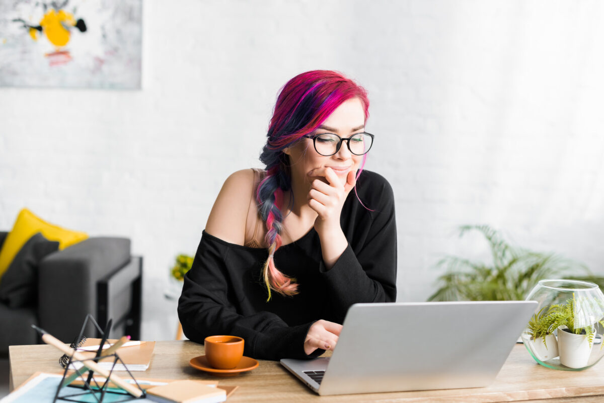 hipster girl with colorful hair sitting behind table and looking at laptop pensively